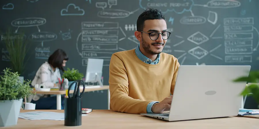 A smiling young professional wearing glasses typing on a laptop at a desk with a complex business diagram drawn on a chalkboard in the background