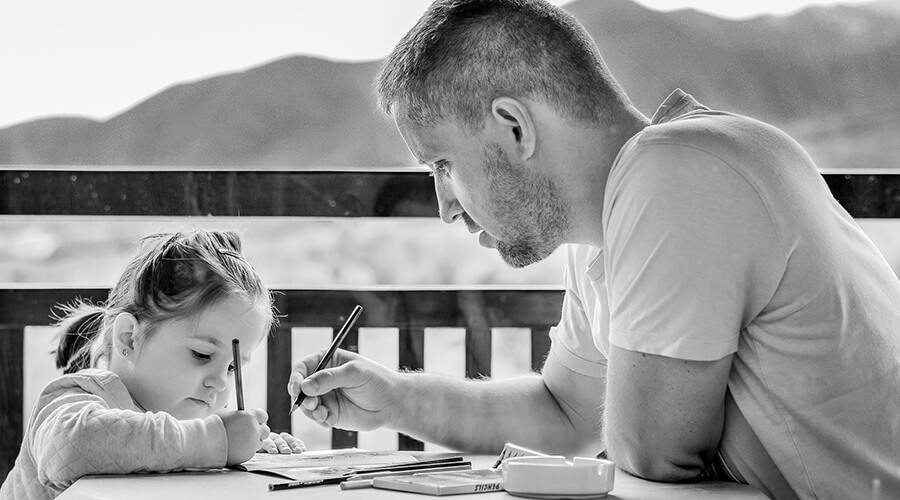 A black and white photo of a man and a young child sitting at a table outdoors. The man is holding a pencil and leaning in to help the child, who is focused on drawing in a book.