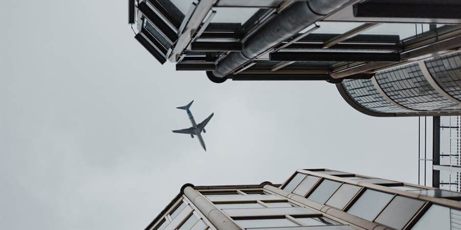 Low-angle view of an airplane flying through a narrow gap between modern city buildings under a grey sky.