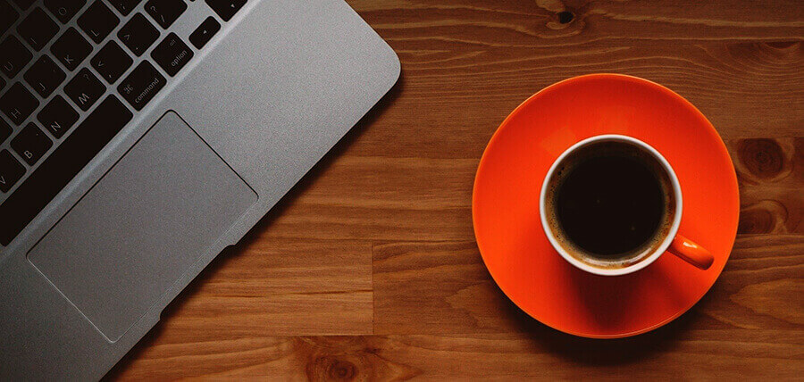 An orange cup of coffee next to a laptop on a wooden table.