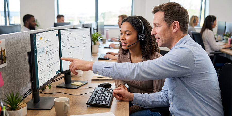 An office worker wearing a headset looks on as her male colleague points at something on one of their two computer monitors at a shared desk. They are focused and collaborating.