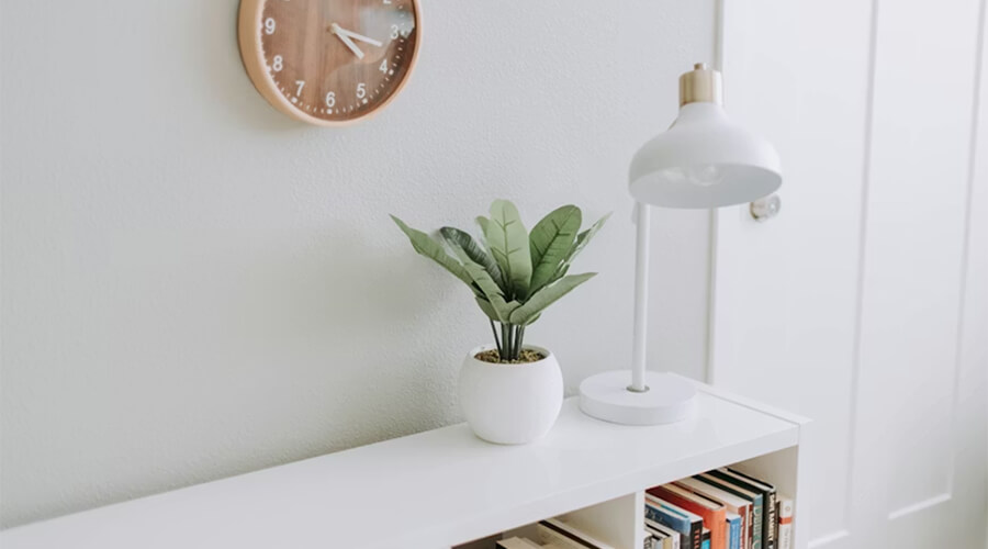 A minimalist and clean white sideboard featuring a small potted green plant, a white desk lamp, and a shelf of neatly organized books. A wooden round clock hangs on the plain white wall above.