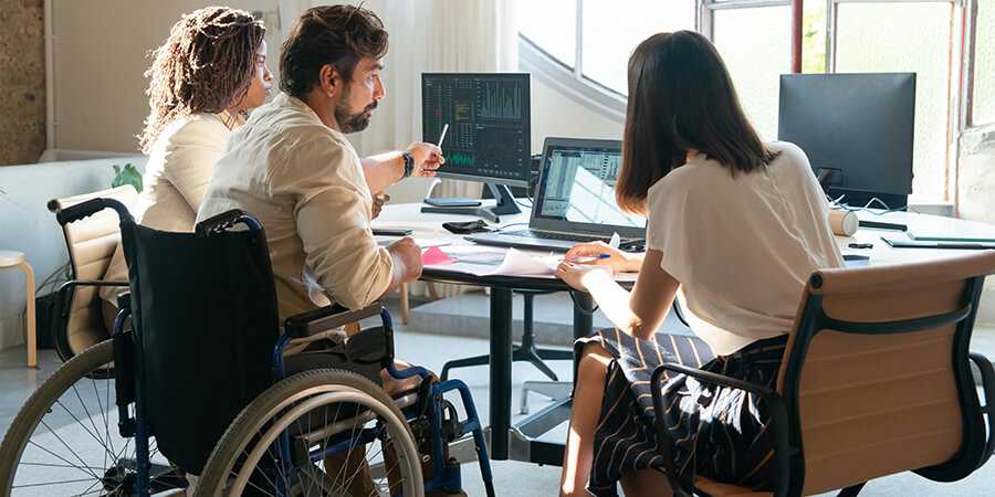 Colleagues in an inclusive office, including a man in a wheelchair pointing to data on a monitor, collaborate around a desk.