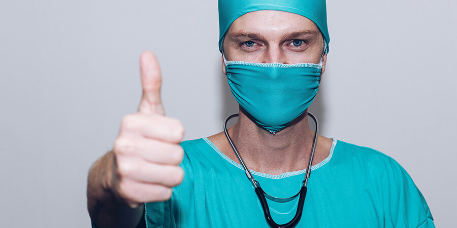 Close up of a medical professional wearing blue scrubs and a mask giving a thumbs up.