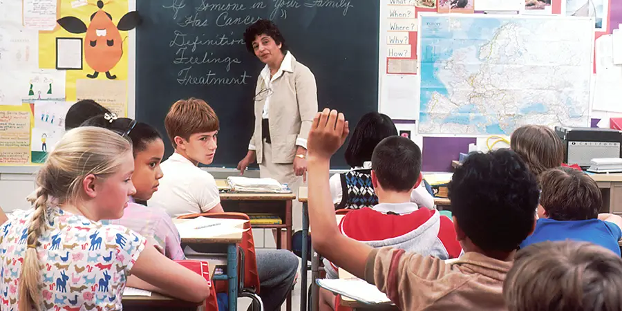 A teacher presents to a diverse classroom of elementary students, with one student raising their hand.