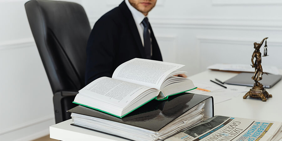 A close-up of a white desk featuring an open thick law book on top of a black binder, with a Lady Justice statue and a person in a suit partially visible in the background.