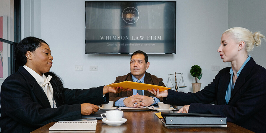 Three professionals in a meeting at Whimson Law Firm; two women are exchanging a large yellow envelope across a wooden table while a man watches from the center.
