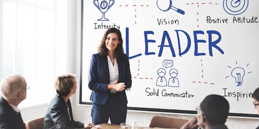 A woman in a business suit presents to her team in front of a whiteboard with the word 'LEADER' and related concepts written on it.