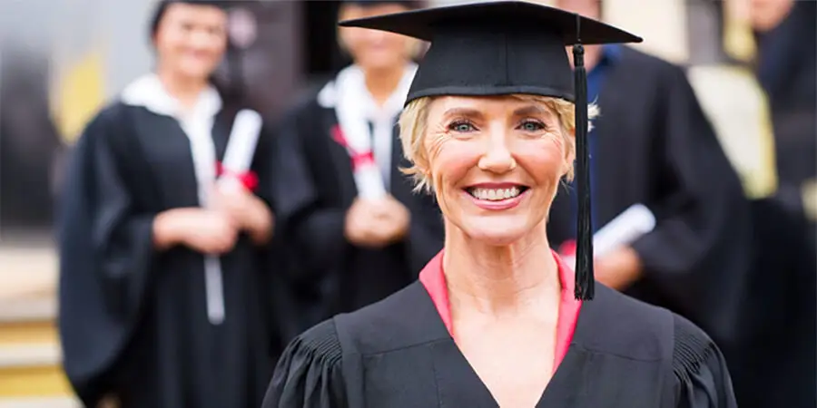 A smiling mature woman wearing a black graduation cap and gown, with other graduates blurred in the background.