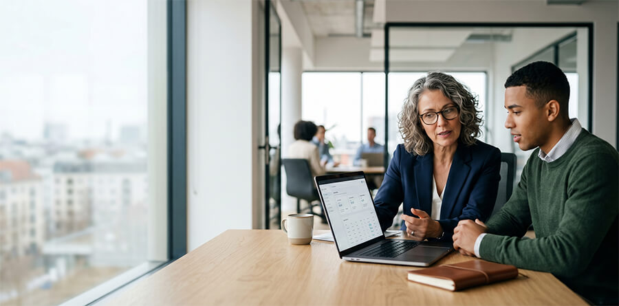 A female professional and a younger male colleague focused on a shared laptop, engaged in an authentic conversation at a wooden desk in an office setting.