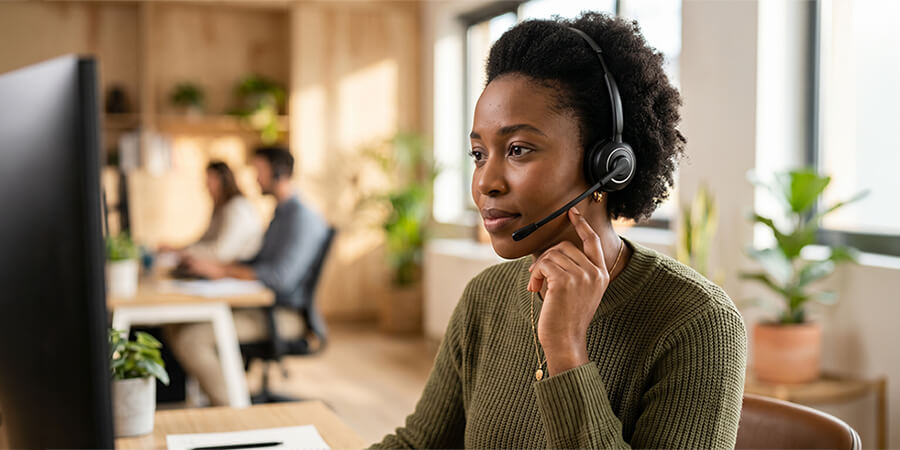 A portrait of a woman wearing a professional headset with a microphone while looking at a computer monitor in a modern office setting.