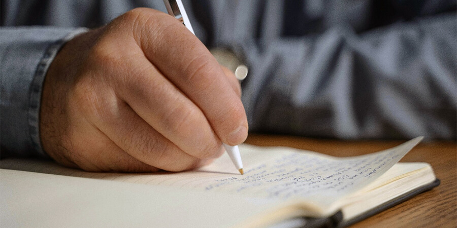 A close-up shot of a person's hand holding a white pen, writing in a lined notebook on a wooden desk.