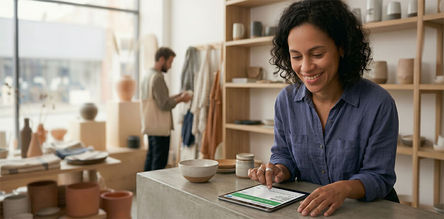 Business owner smiling while checking online sales on a tablet at shop counter, boutique in background.