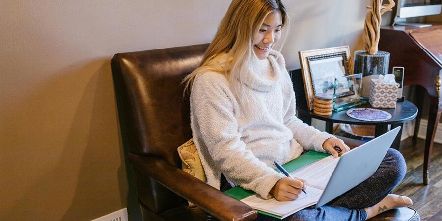 A smiling woman sitting comfortably in a leather armchair, working on a laptop and taking notes in a green folder.