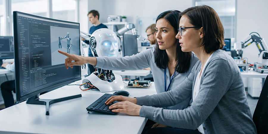 Two female engineers and a humanoid robot review code on a computer screen in a robotics laboratory.