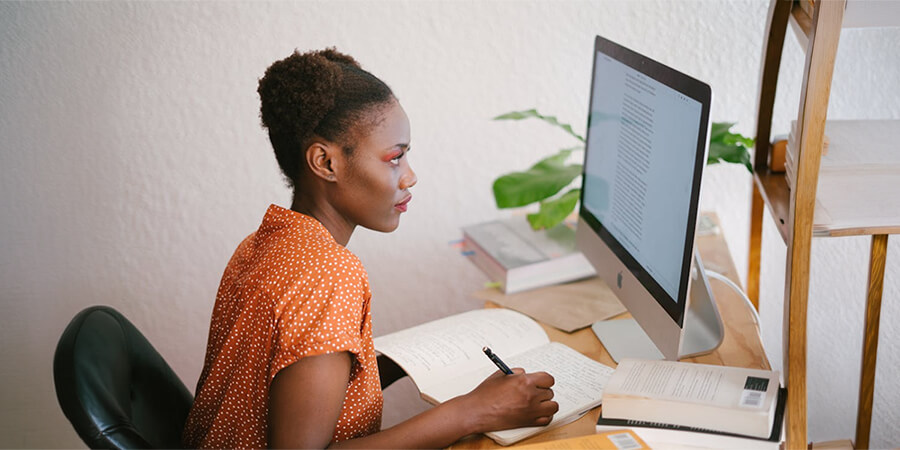 A woman sitting at a desk, focused on a computer screen while writing notes in an open book.