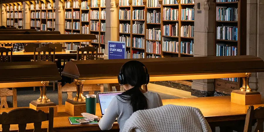 A person wearing headphones works on a laptop at a large table inside a grand library with many bookshelves.