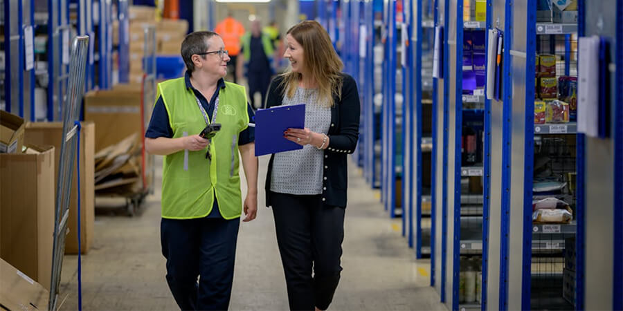 Two women walking through a warehouse aisle and talking; one is wearing a high-visibility safety vest and holding a scanner, while the other holds a blue clipboard.