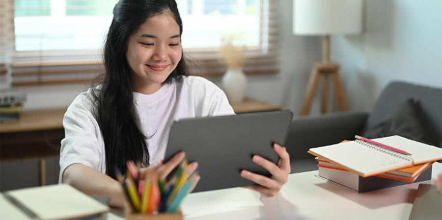 A smiling young woman with long dark hair sits at a desk in a bright room, holding a black tablet. The desk is organized with a cup of colored pencils, notebooks, and a stack of textbooks.