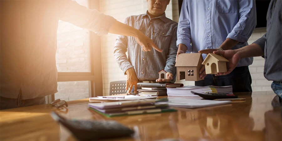 Small wooden house model standing on a table next to stacks of coins and a calculator.