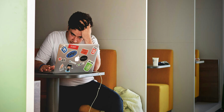 A person sitting in a small office booth, looking down at a laptop screen with one hand on their forehead. The laptop lid is covered in various tech-related stickers including PHP, HTML5, and GitHub.