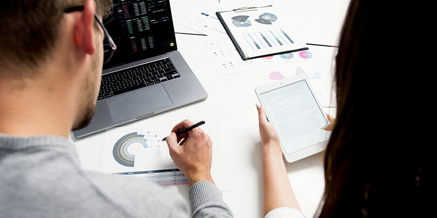 An over-the-shoulder view of two people working at a white desk, analyzing printed charts, a laptop, and a tablet.