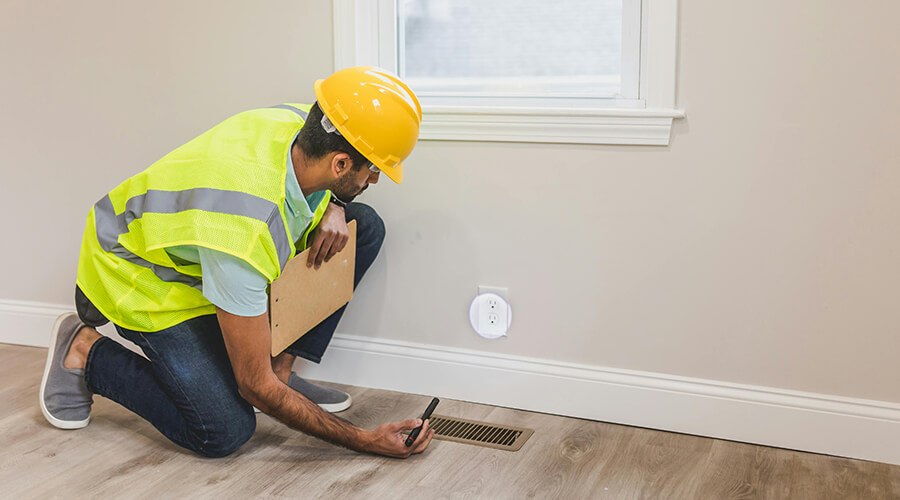 A building inspector in a hard hat and safety vest kneeling to examine an electrical outlet with a flashlight.
